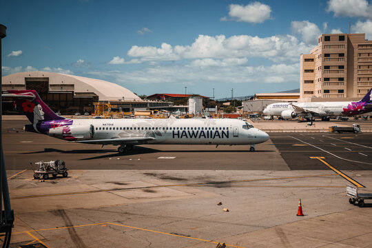 Commercial passenger aircraft fleet on the ramp. Hawaiian Airlines Boeing 717 regional jet and Boeing 787 Dreamliner twin-engine widebody plane at airport terminal under blue sky.