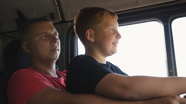 Close up of smiling ginger child holds the big steering wheel sitting on lap of father and learning to drive the truck. Happy dad teaches his red-haired kid to operating a car at empty road