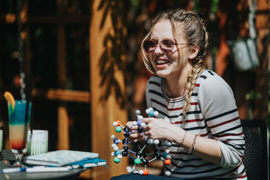 A teenage girl with braided hair and braces builds a colorful molecular model. A school friend smiles while assembling the educational model at an outdoor cafe.