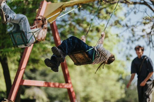 Two friends swing high on a park swing set, smiling and enjoying the moment. The scene shows playful outdoor fun with young people on swings in a leafy playground.