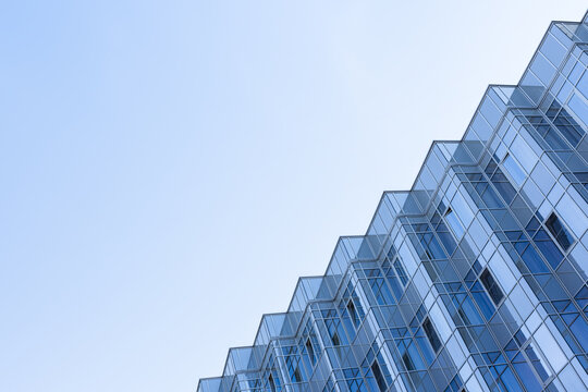 Modern glass office building facade photographed from an upward angle, showcasing repeating geometric patterns, reflective blue windows and minimalist corporate architecture against a clear sky