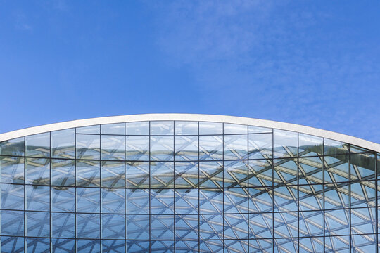 Curved glass facade displays repeating geometric patterns and reflective surfaces against a clear blue sky, highlighting modern architecture, transparency, symmetry and contemporary structural design