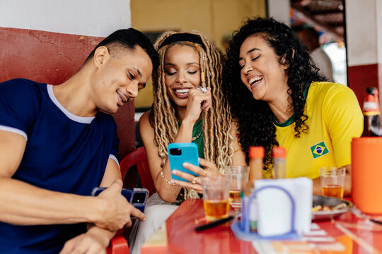 Brazilian friends smiling while looking at smartphone together at a casual bar, sharing content and enjoying a relaxed social moment.