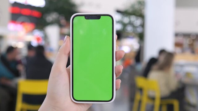 Hand holds smartphone with green screen. Blurred background shows indoor cafe setting. Yellow chairs and people visible. Focus on phone, not on background details