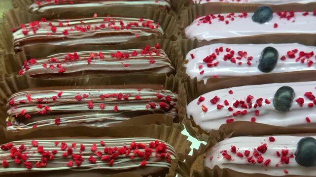 Close-up assorted eclairs on display in coffee shop case, rows of chocolate and vanilla eclairs topped with red sprinkles and fresh