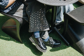 Fototapeta premium Two school friends sit with crossed legs under a round outdoor table, showing black shoes and denim. Casual students relax on patio chairs on artificial grass in a sunny outdoor setting.