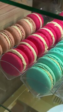Colorful macarons coffee shop window close-up, rows of teal pink orange and brown shells arranged on clear tray inside warm lit