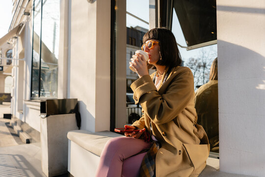 Young woman in a beige coat sipping coffee outside a city cafe while checking her phone