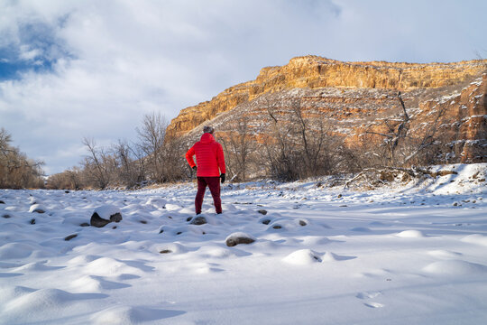 hiker in down jacket on a shore of frozen Poudre River with monumental sandstone cliff at Colorado foothills of Rocky Mountains near Fort Collins
