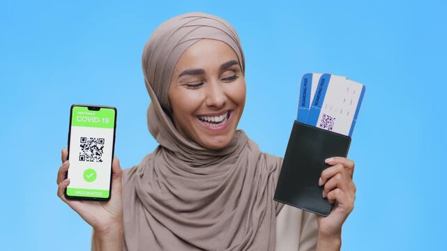 A woman smiles widely while holding a smartphone displaying a COVID-19 vaccination QR code. She also holds a wallet with travel tickets. The background is bright blue.