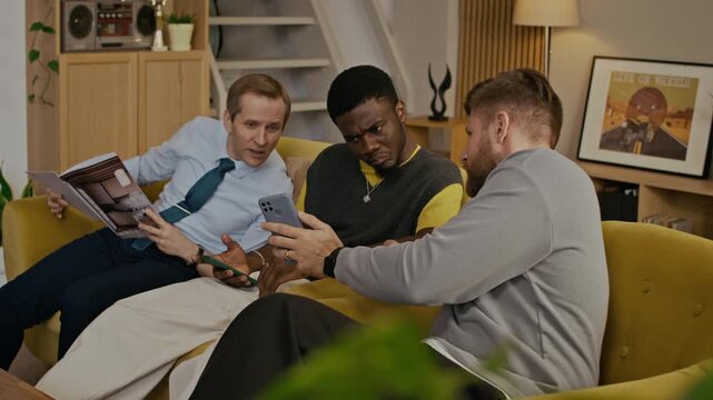 Handheld shot of three biracial best friends resting on sofa, reading fashion magazine and checking social networks on smartphones