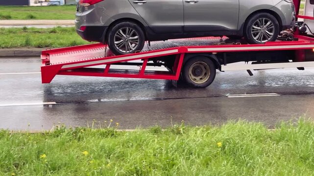 Close-up passenger car on red flatbed tow truck, spring roadside scene with wet pavement and puddles, green grassy verge dotted