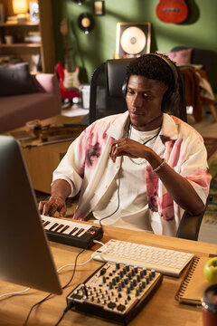 Vertical shot of young Black man producing music on computer, wearing headphones and using midi keyboard in home studio. Supporting audio production, creator economy, remote work marketing