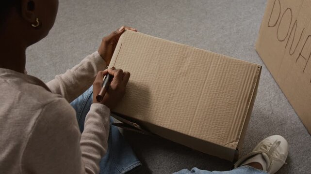 Over the shoulder view of African American girl writing word donation with black marker on cardboard box while sitting on carpeted floor