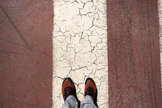 Overhead view of feet in orange sneakers standing on cracked white street stripe between brown pavement. Urban texture and weathered road markings. For journey and decision concepts.
