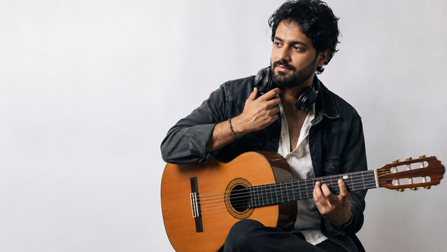 Musician with Guitar and Headphones: A pensive musician with a beard and headphones around his neck sits with a classical guitar, his fingers poised on the fretboard, in a thoughtful pose.