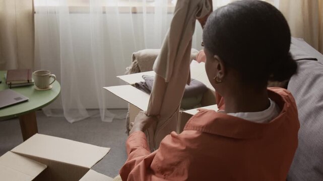 Tilt down view of Black gen Z girl sorting clothing items and placing them into box while decluttering her wardrobe in living room