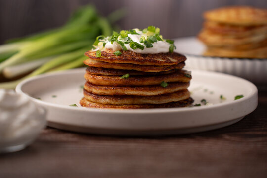 close-up of a warm stack of golden-brown savory pancakes on a white ceramic plate, topped with creamy sour cream and chopped green onions, set on a rustic wooden table with soft natural light