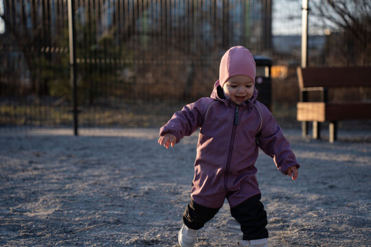 smiling toddler in a purple one-piece snowsuit and pink beanie toddling across a sandy urban playground at golden hour, warm side light casting long shadows by a bench and metal fence, softfocus