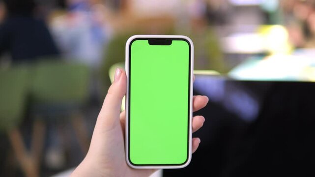 Hand holds smartphone with green screen. Background shows blurred cafe seating. Phone has rounded corners and notch at top. Focus remains on phone throughout clip. No animals or plants visible