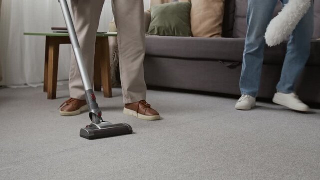 Low section view of unrecognisable man wearing grey pants and brown shoes cleaning carpet with upright vacuum, anonymous woman with feather duster grabbing clothing item from couch