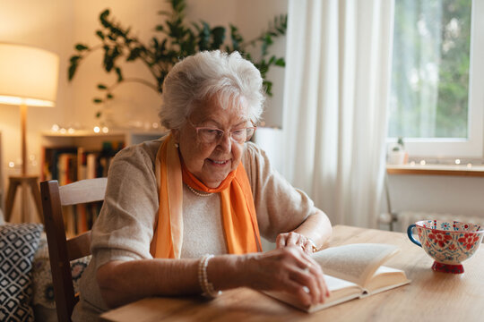 Side view of elderly lady reading a book, sitting at the kitchen table and sipping tea.