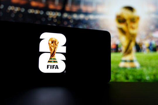 Official FIFA World Cup 2026 brand logo displayed on a digital screen alongside a blurred trophy background, concept of international soccer tournament and football championship merchandise in the USA
