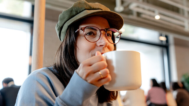 Woman in cafe sipping coffee, wearing glasses and cap