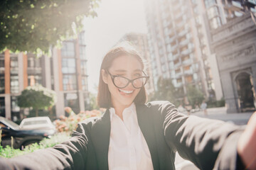 Fototapeta premium Smiling businesswoman taking a selfie in an urban street wearing glasses and professional blazer on a sunny day