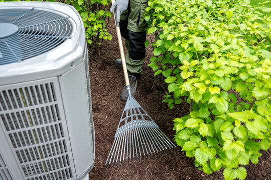 Landscaper raking fresh mulch around bushes in residential backyard close up to air condition. yard maintenance work in suburban 