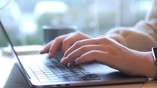 Hands type on laptop keyboard. Soft natural light from window. Background blurred with coffee cup. Person wears light sweater. Watch on wrist. Focused work scene. Calm indoor setting