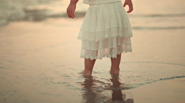 Low angle cropped view of toddler girl in white dress walking barefoot on wet sand at beach during sunset gentle sea waves and warm golden hour light carefree childhood vacation concept