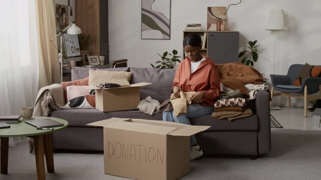 Black young woman sorting clothing items and putting them into cardboard box while preparing for donation sitting on grey couch in living room