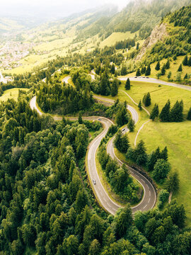 Travelers navigate a winding asphalt road through lush greenery in the Alps. The aerial view captures the curves of the serpentine path and surrounding hills during a sunny day.