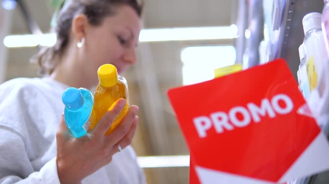 Young caucasian woman shopping in a store, comparing two cosmetic bottles and smelling a product from the shelf near a red promo sign, making a difficult choice while buying beauty items
