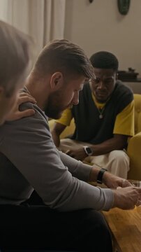 Vertical shot of biracial friends supporting best friend, patting his shoulder and trying to cheer him up while he talking about life drama