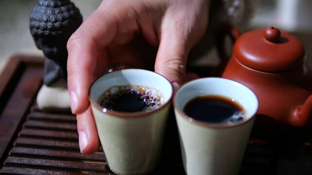 Close up view of a person's hand picking up a small cup of hot brewed puerh tea during a traditional chinese gongfu ceremony, with a clay teapot and a buddha statue in the background