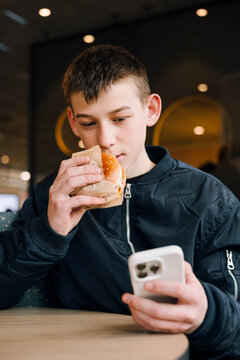 Teenage boy eating burger while checking smartphone indoors