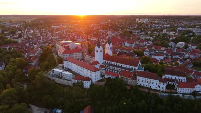 A drone panorama over Freising, Bavaria, Germany, shows the cathedral hill, St. George Church, and the historic city center framed by red rooftops and soft sunset colors.