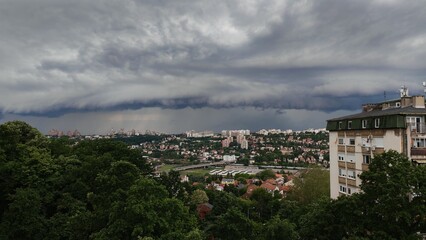 Obraz premium A dark storm cloud hanging over a city with buildings and forest in the background. Dramatic weather scene with contrast between urban area and nature.