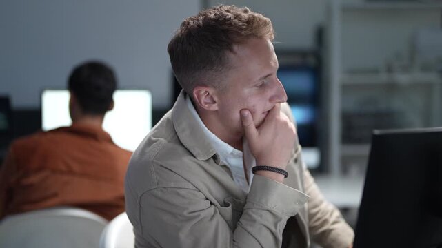 A software developer focusing on programming desktop computer monitors in a corporate office workspace while a colleague works in the background.