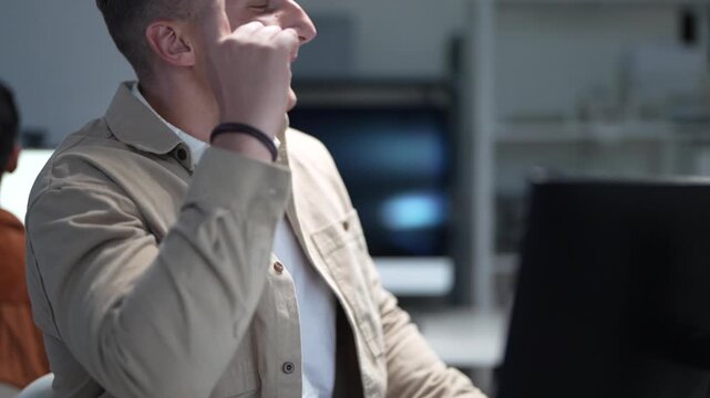 A software developer focusing on programming desktop computer monitors in a corporate office workspace while a colleague works in the background.