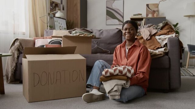 Portrait of African American young woman with pile of sweaters looking at camera with smile while preparing clothes for donation sitting on carpeted floor in living room