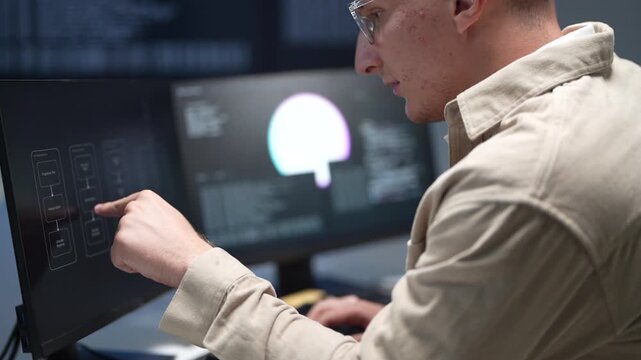 A software developer focusing on programming desktop computer monitors in a corporate office workspace while a colleague works in the background.