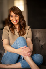 Fototapeta premium Smiling mature woman sitting cross-legged on a comfortable sofa, looking at camera