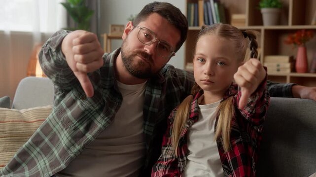 Dissatisfied Caucasian man dad little girl daughter showing thumb down dislike frustrated kid child schoolgirl at home male parent father guy family negative bad gesture rejection disagree disapproval