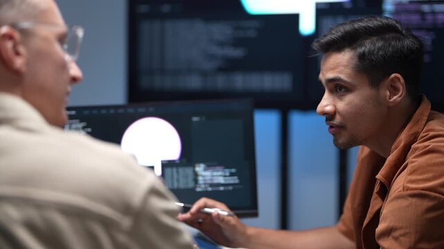 Close up of software developers sitting at an office desk to discuss machine learning code scripts and system architecture changes on a desktop monitor.