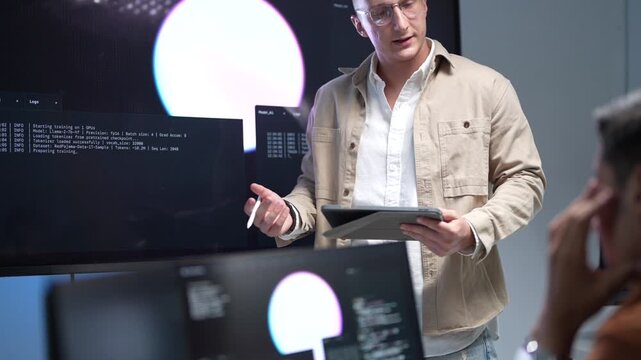 A senior IT specialist wearing glasses and holding a tablet while presenting machine learning workflows and artificial intelligence algorithms on a large screen.