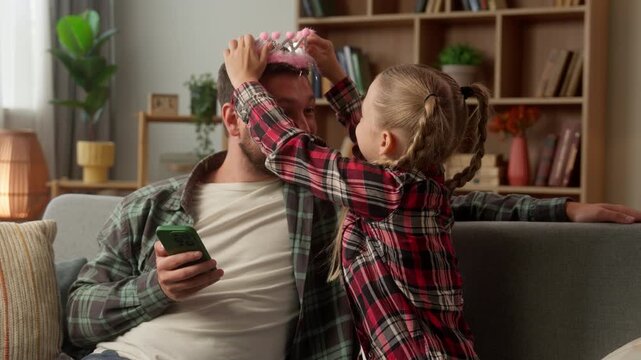 Happy Caucasian family man dad little girl daughter putting pink crown laughing guy using mobile phone child distract parent schoolgirl father funny kid game together male at home parenthood enjoying