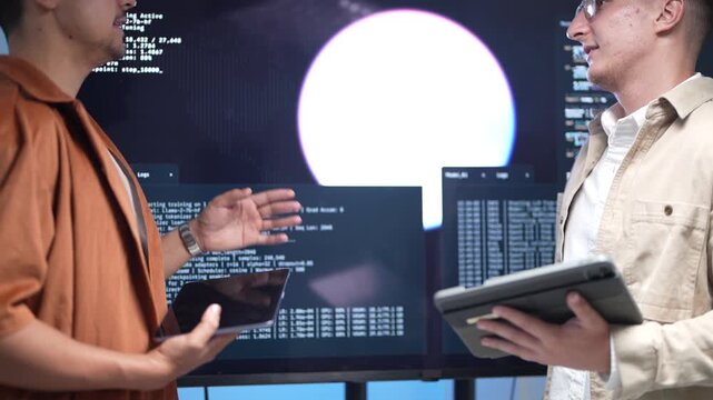 wo cheerful IT specialists holding digital tablets and discussing artificial intelligence data structures and machine learning scripts in front of big screens.